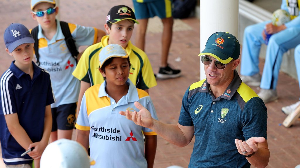 Cricket coach in a blue and yellow shirt talks to a group of young players in sporty uniforms outdoors.
